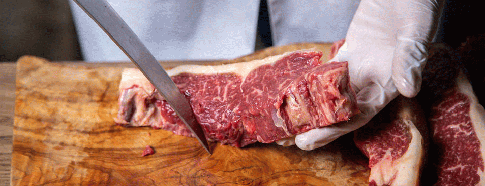 Butcher Cutting Meat in White Gloves Holding Big Knife on the Wooden Desk